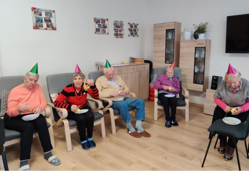 Seniorzy siedzą na fotelach a na głowach maja kolorowe czapeczki